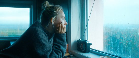 Szenenbild aus dem Drama THE OUTRUN: Porträt einer jungen Frau, die an einem Fenster steht und nach draußen in eine verregnete Landschaft schaut. Sie hält eine Zugarette in der Hand. Das Bild ist in blau-grauen Farbtönen gehalten.