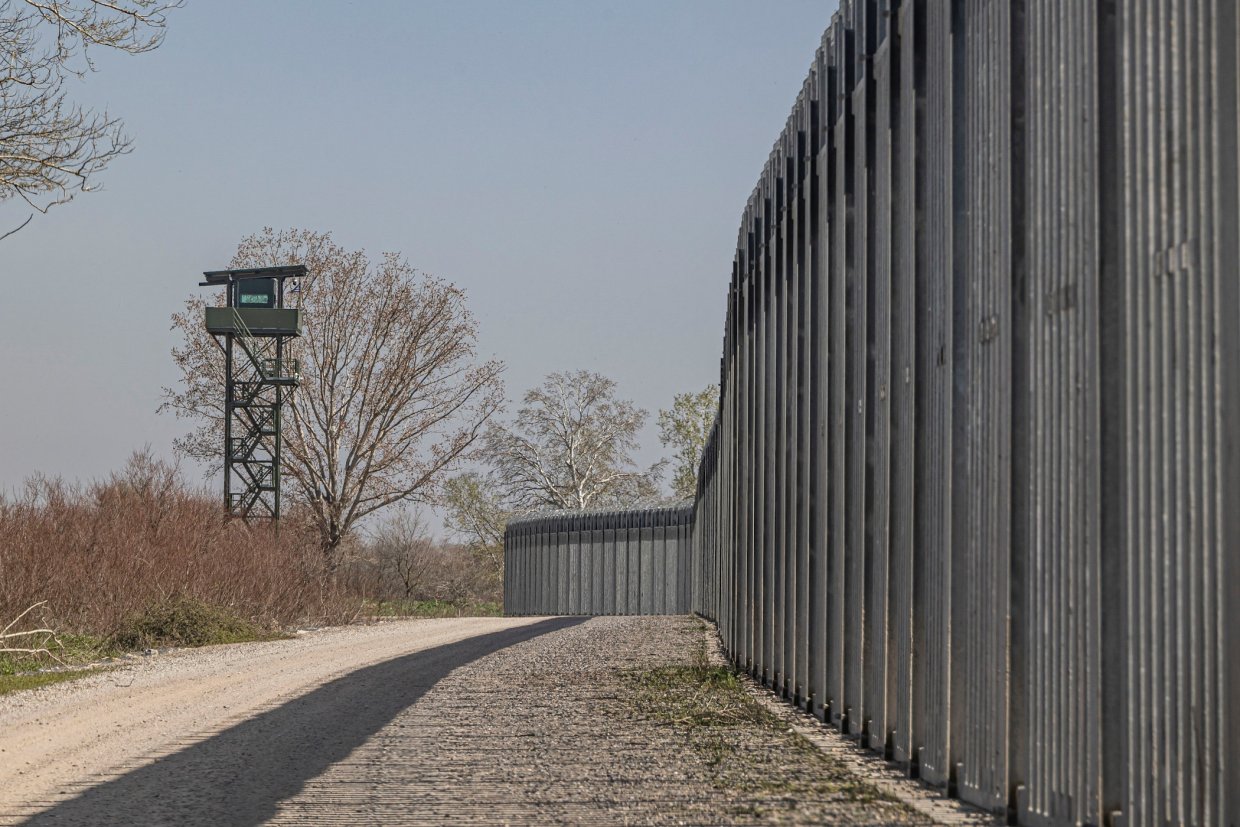 Farbfoto: Eine lange graue Mauer nimmt die rechte Seite des Bildes ein. Mittig ist ein Schotterweg zu erkennen und links davon sieht man einen Grenzturm sowie Bäume und Büsche.