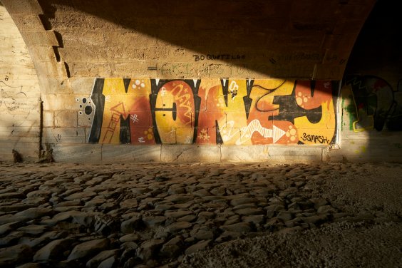 Es ist die Unterführung einer Brücke zu sehen. Auf der Mauer wurde in rot-gelber Schrift ein Graffiti gesprüht, welches das Wort "Money" darstellt.